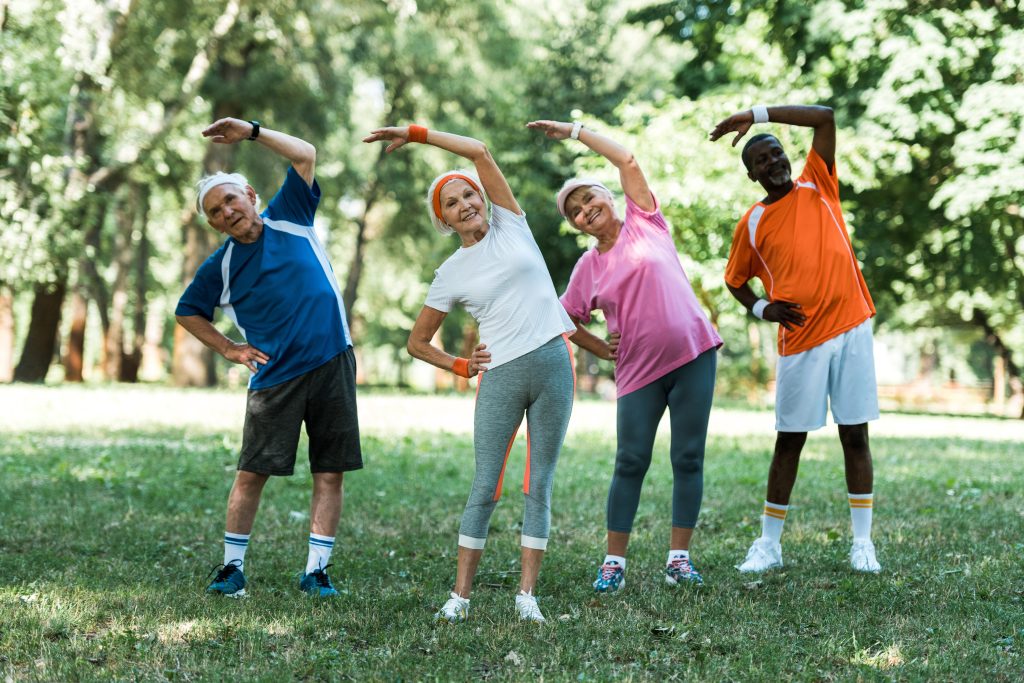 cheerful multicultural retired men and women standing with hands on hips while doing stretching exercise on grass